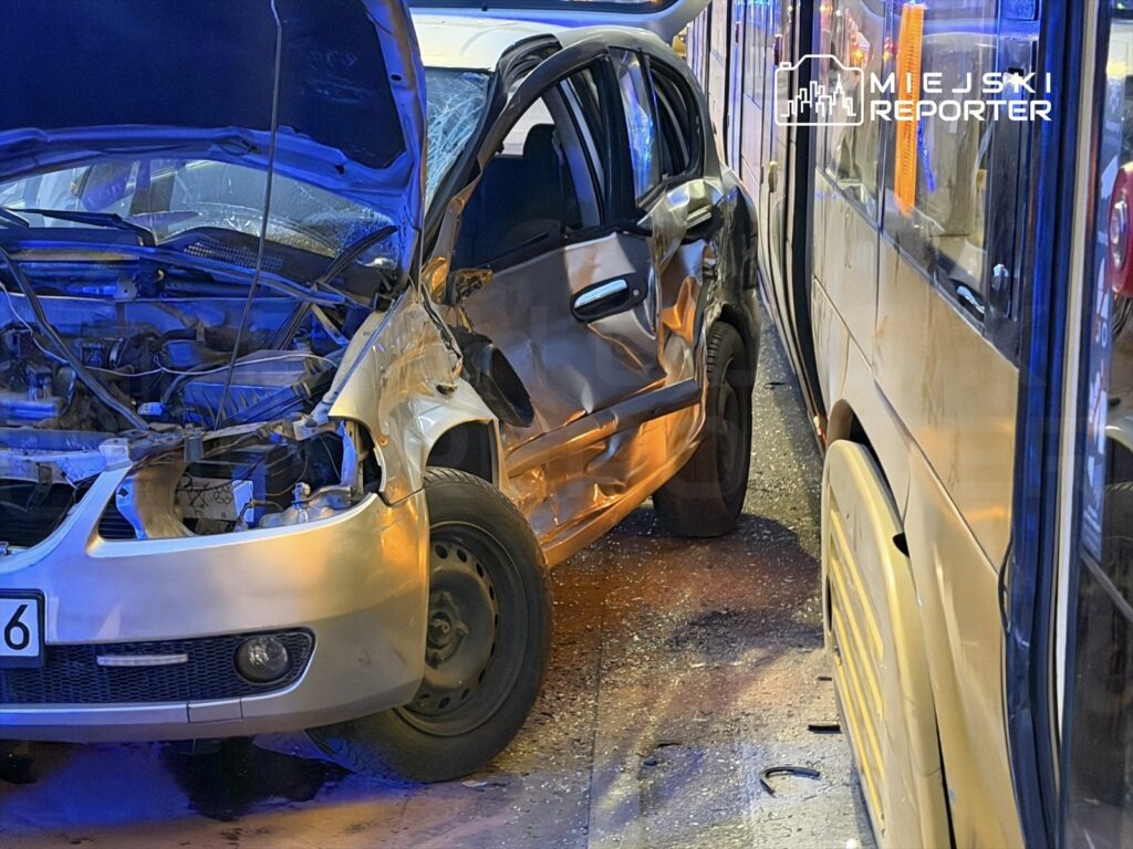 Uszkodzony samochód osobowy z wgniecioną stroną stoi obok autobusu w miejskim otoczeniu, oświetlony niebieskimi i pomarańczowymi światłami.