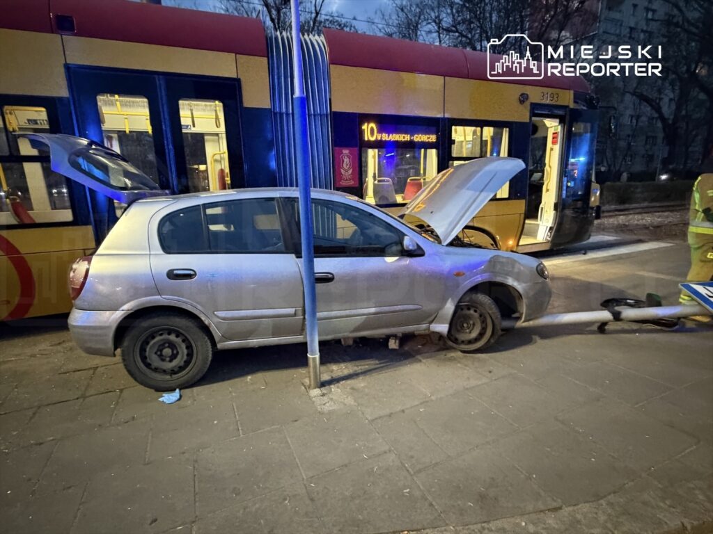 Uszkodzony srebrny samochód osobowy zaparkowany obok przystanku tramwajowego, z otwartą maską i uniesionym tylnym kołem.