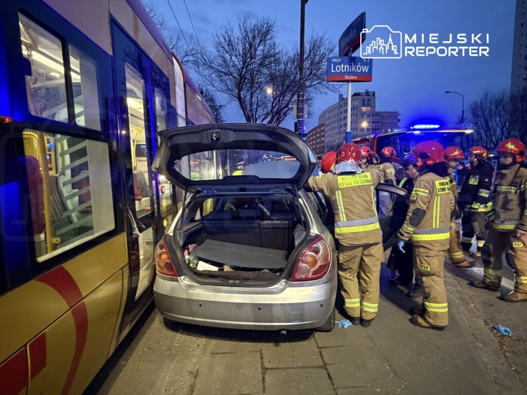 Zespół strażaków w kaskach pracuje przy uszkodzonym samochodzie, obok przystanku tramwajowego w miejskim otoczeniu.
