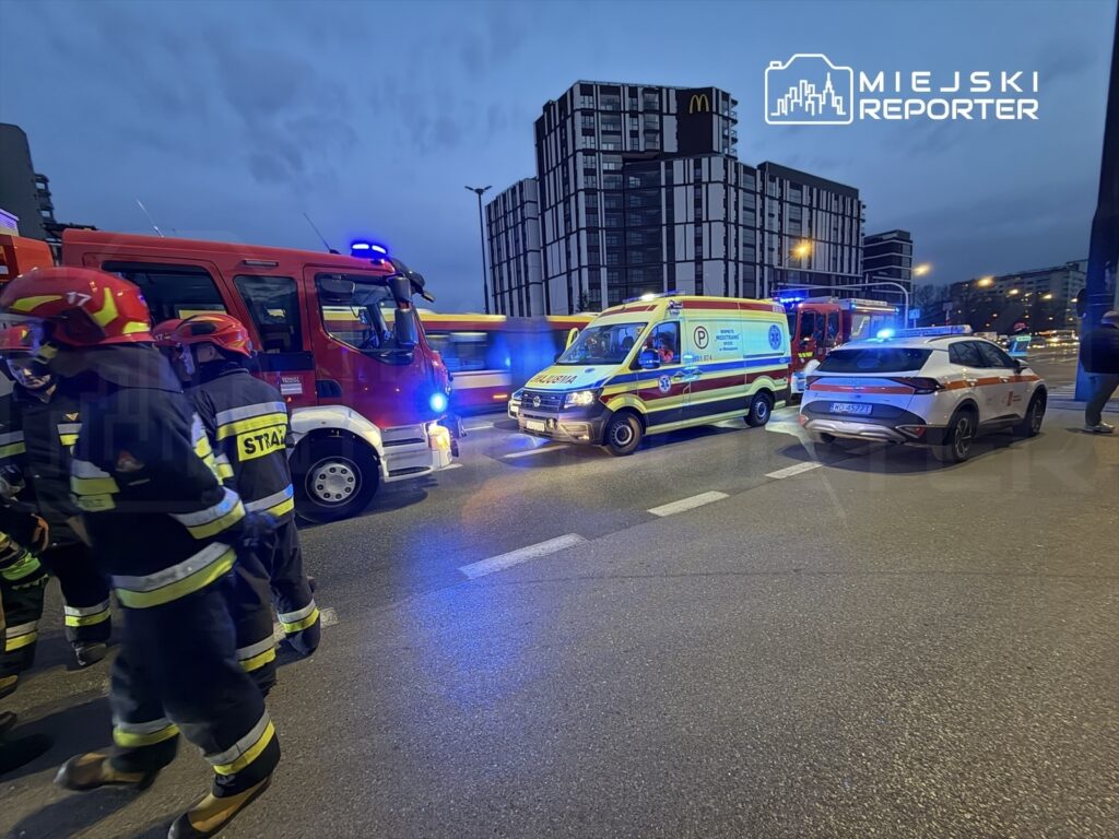Grupa strażaków w mundurach rozmawia obok wozu strażackiego i karetki na miejskiej ulicy wieczorem.