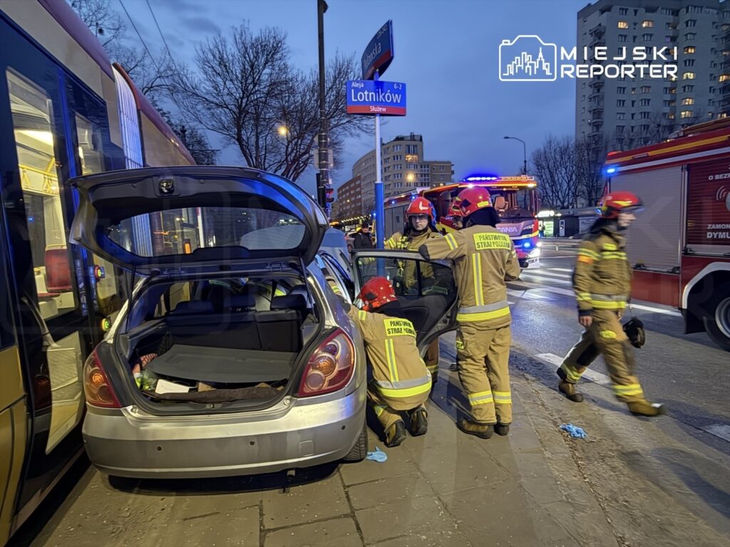 Strażacy w uniformach pracują przy uszkodzonym samochodzie na ulicy, obok przejeżdżającego tramwaju.