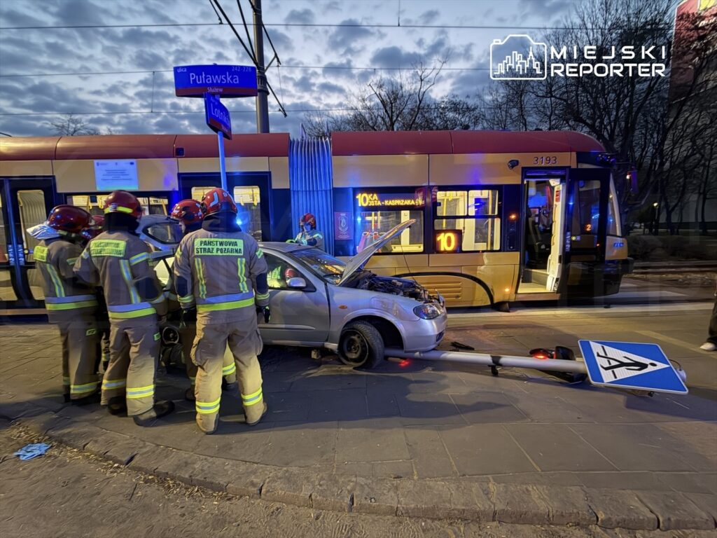 Grupa strażaków w mundurach z odblaskami udziela pomocy przy rozbitym samochodzie obok tramwaju.