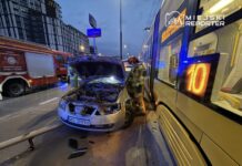 Strażak sprawdzający uszkodzony samochód osobowy z otwartą maską obok przystanku autobusowego w miejskim otoczeniu.