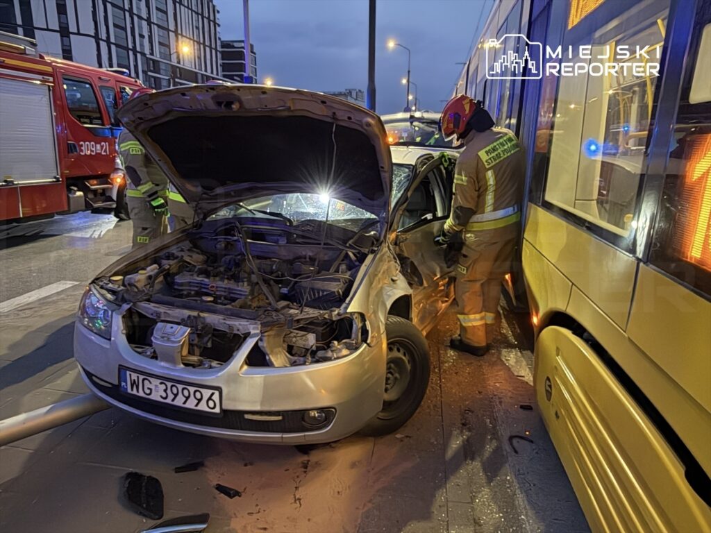 W nocy strażak w specjalnym ubraniu sprawdza uszkodzony samochód osobowy, który zderzył się z tramwajem.