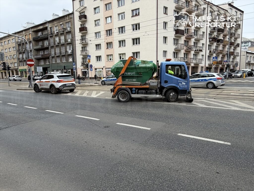 Śmieciarka z zielonym kontenerem z logo "PERFECT" jedzie ulicą obok białego samochodu służb miejskich.