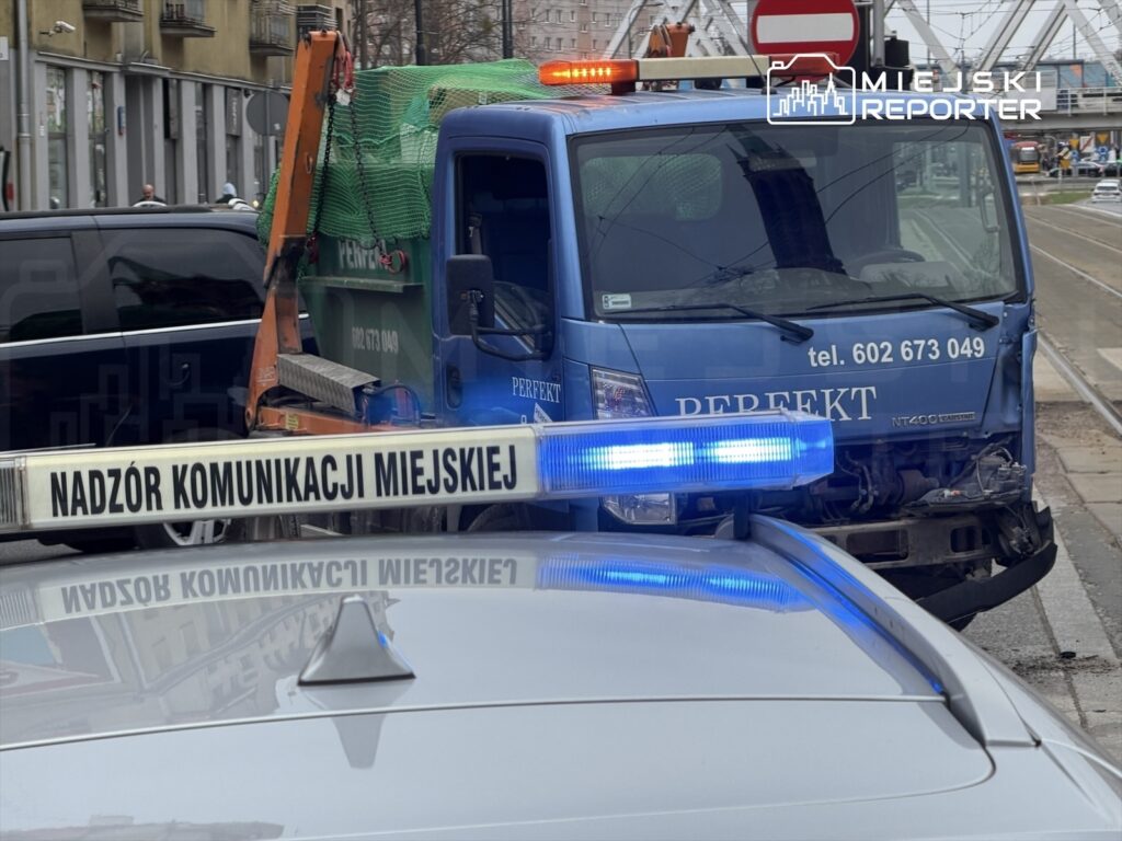 Kolizja drogowa z udziałem niebieskiego pojazdu dostawczego z uszkodzoną przednią częścią, obok stoi samochód z sygnalizacją.