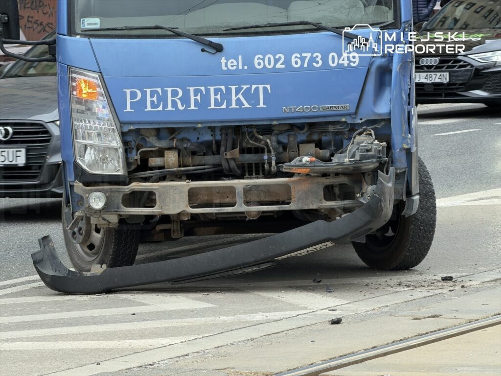 Uszkodzony niebieski furgon z widoczną uszkodzoną przednią częścią stoi na drodze, obok przejazdu tramwajowego.