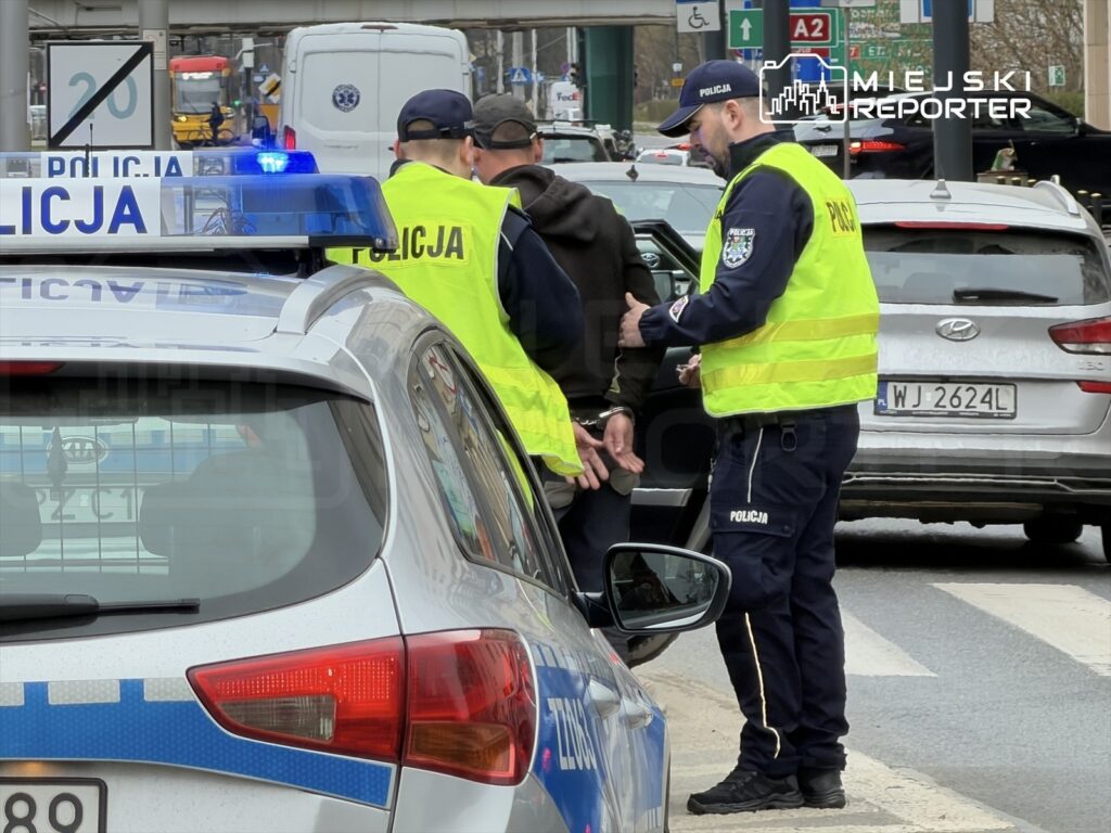 Dwaj policjanci w kamizelkach odblaskowych rozmawiają z mężczyzną przy radiowozie na ruchliwej ulicy.