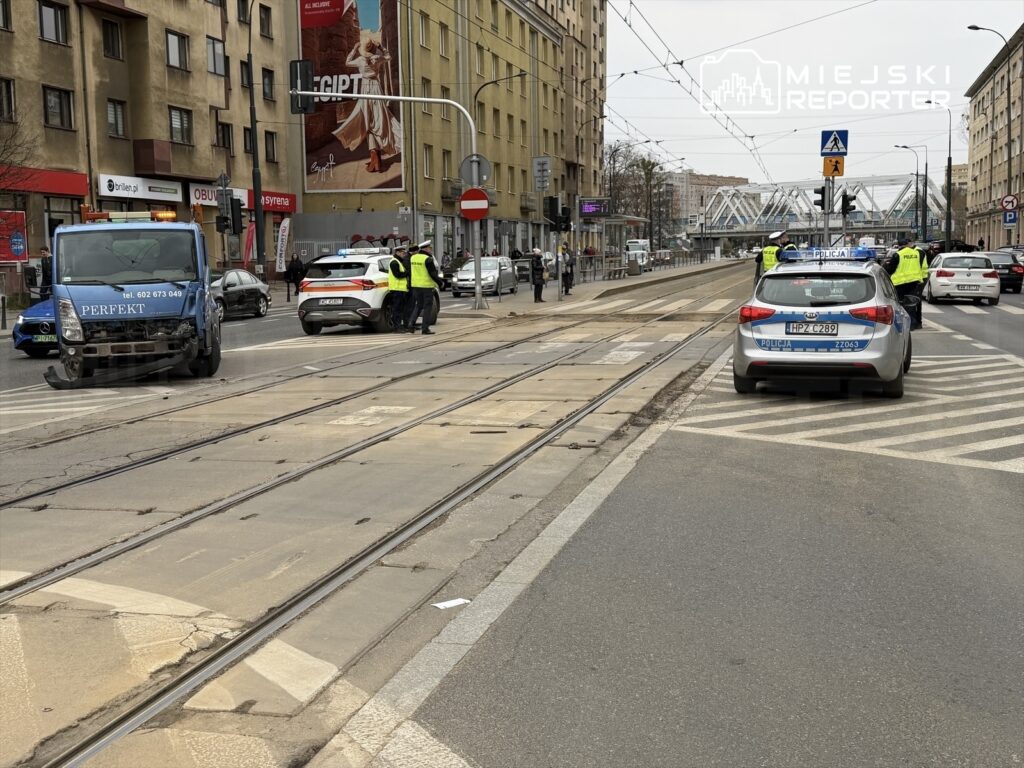 Na skrzyżowaniu ulicy stoją radiowozy policji, a funkcjonariusze rozmawiają z kierowcami. W tle widać zniszczony samochód dostawczy.