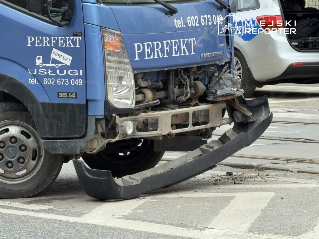 Uszkodzony niebieski samochód ciężarowy z oderwaną przednią zderzakiem stoi na ulicy obok torów tramwajowych.