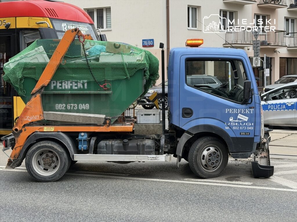 Mały niebieski samochód dostawczy z kontenerem na odpady, zaparkowany na ulicy obok przystanku autobusowego.