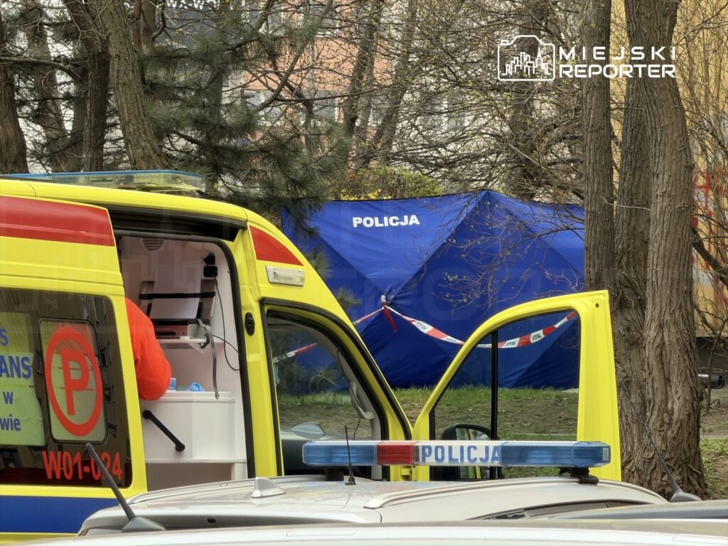 Karetka pogotowia z otwartymi drzwiami, w tle niebieski namiot policyjny z napisem „POLICJA” i oznaczeniami taśmy zabezpieczającej.