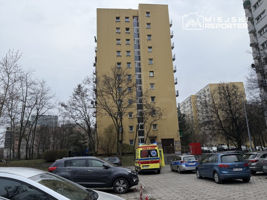 Karetka pogotowia i radiowóz stoją na parkingu przed żółtym blokiem mieszkalnym w szarej, pochmurnej scenerii.