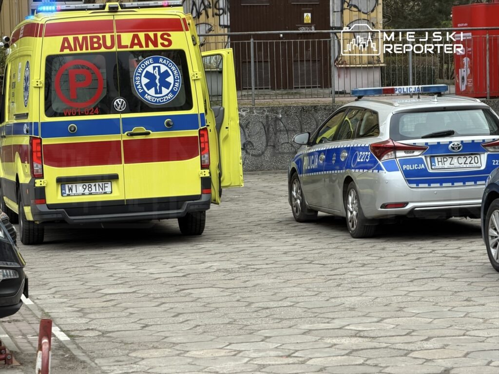 Karetka pogotowia i radiowóz policyjny zaparkowane na wybrukowanej ulicy, w tle widoczne graffiti na murze.