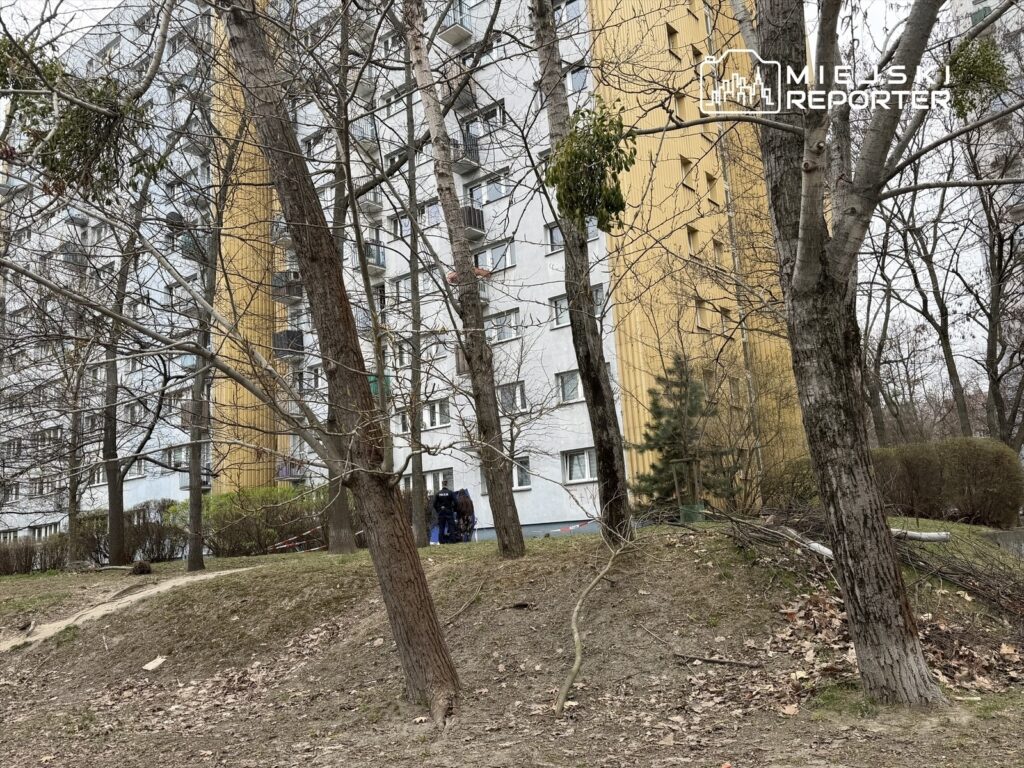 Mężczyzna z wózkiem spacerowym przechodzi obok nowoczesnego budynku mieszkalnego otoczonego nagimi drzewami.