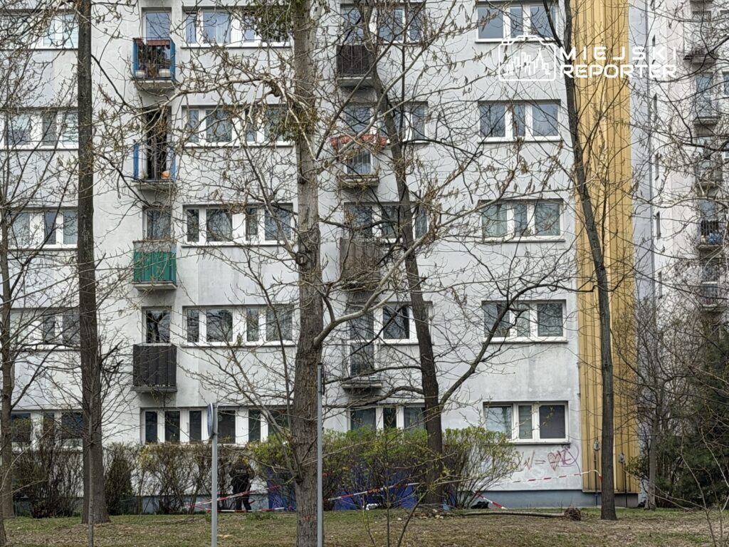 Wielopiętrowy blok mieszkalny z balkonami w różnych kolorach, otoczony nagimi drzewami i zaroślami.