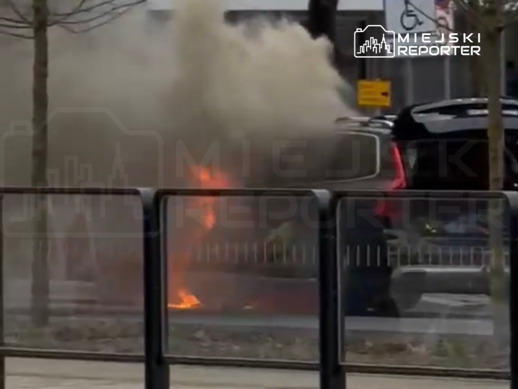 W tle widać dym i ogień wydobywające się z pojazdu zaparkowanego przy ulicy, otoczonego barierkami.