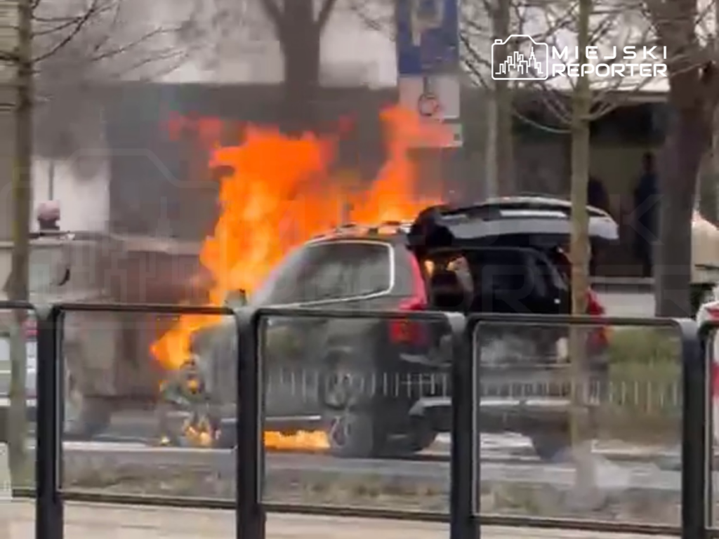 Samochód osobowy płonie na ulicy, otoczony przez dym i ogień, w tle widoczne inne pojazdy oraz przechodnie.