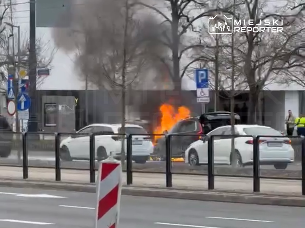 Pojazd zapalił się na ulicy, w tle widoczny dym i ogień, a w pobliżu stoją samochody oraz osoby w mundurach.
