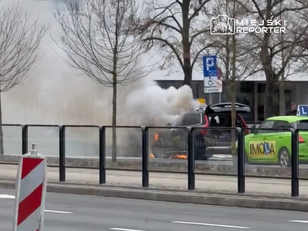 Samochód wydobywa gęsty dym i płomienie na ulicy, obok zaparkowane taksówki i drzewka.