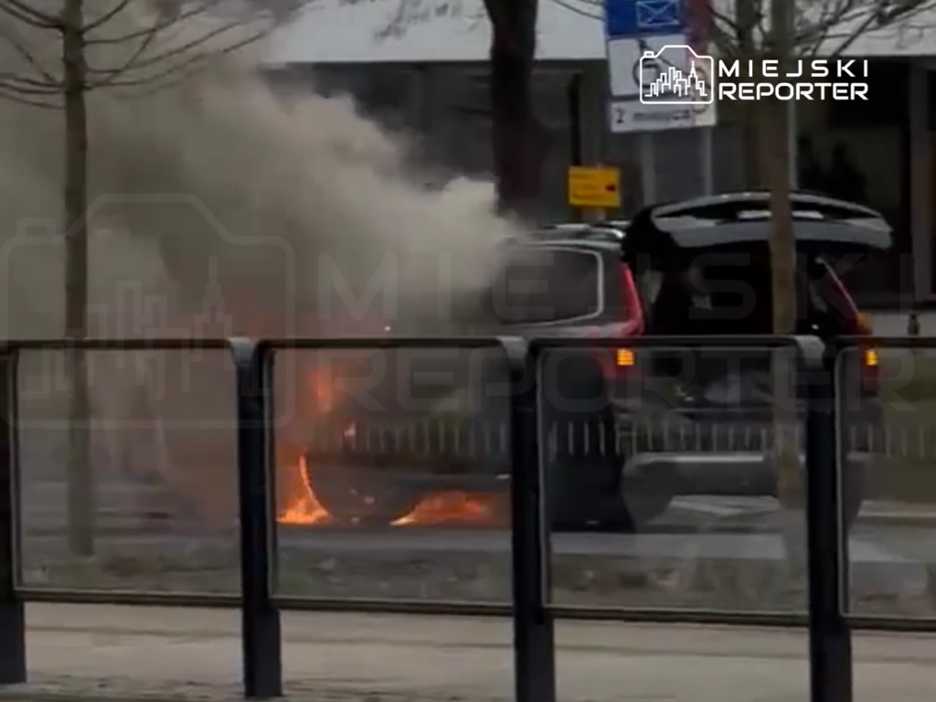 Samochód zaparkowany na ulicy, z którego wydobywa się dym i ogień, otoczony przez barierki.