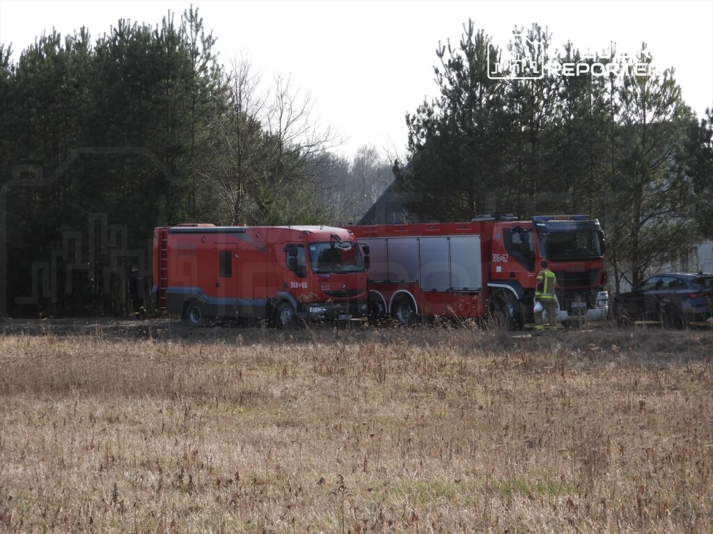 Dwa czerwone wozy strażackie zaparkowane na polnej drodze w otoczeniu drzew i zarośli. Obok wozu stoi strażak w kamizelce odblaskowej.