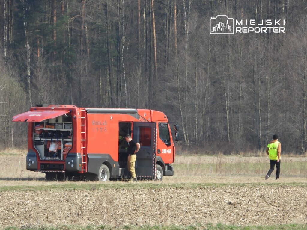 Czerwony pojazd mobilny MobiLab stoi na polu, a mężczyzna w kamizelce odblaskowej idzie w kierunku lasu.