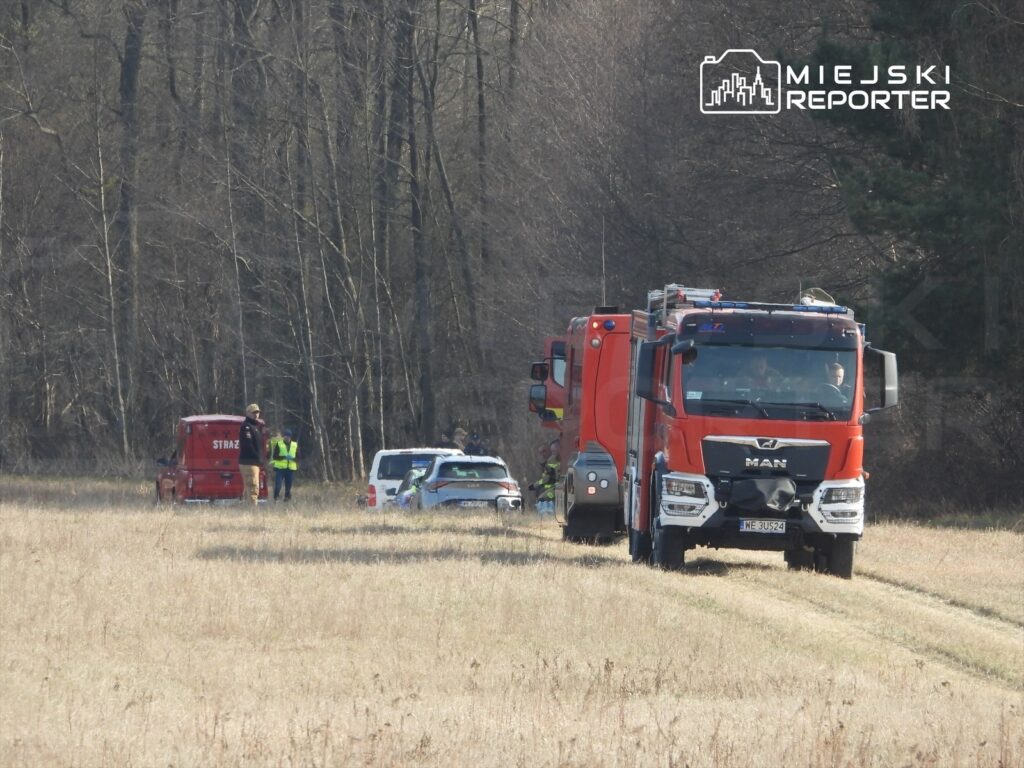Grupa strażaków i policjantów prowadzi działania w terenie, obok wozu strażackiego i radiowozu.