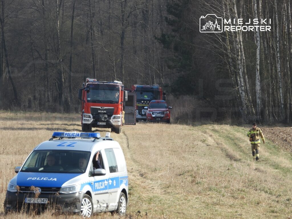 Pojazd policyjny i samochody strażackie poruszają się po polnej drodze, a strażak idzie w kierunku lasu.