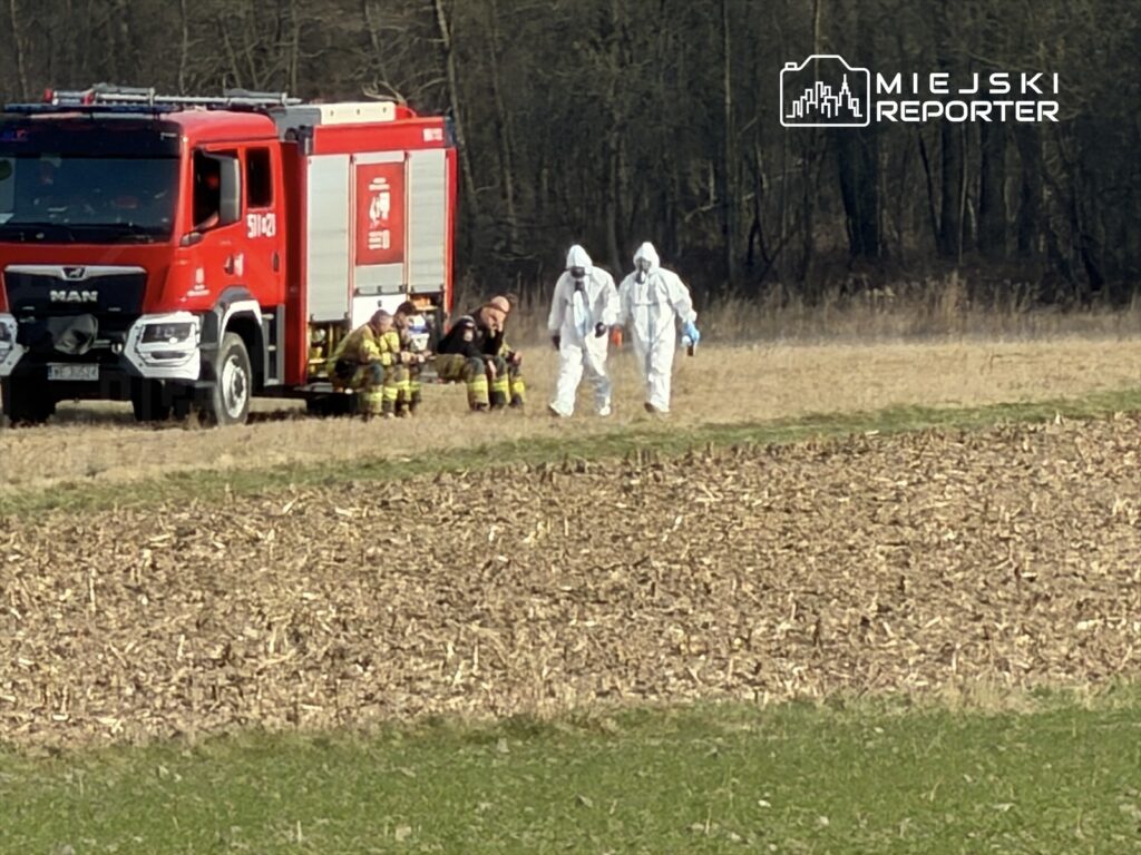Czterej strażacy w mundurach siedzą na ziemi obok wozu strażackiego, podczas gdy dwaj mężczyźni w białych kombinezonach idą w kierunku lasu.