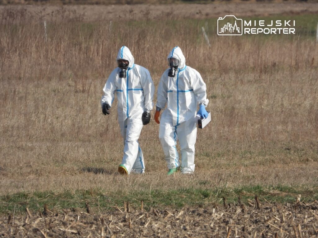 Dwaj mężczyźni w kombinezonach ochronnych i maskach gazowych idą przez pole, trzymając dokumenty i sprzęt.