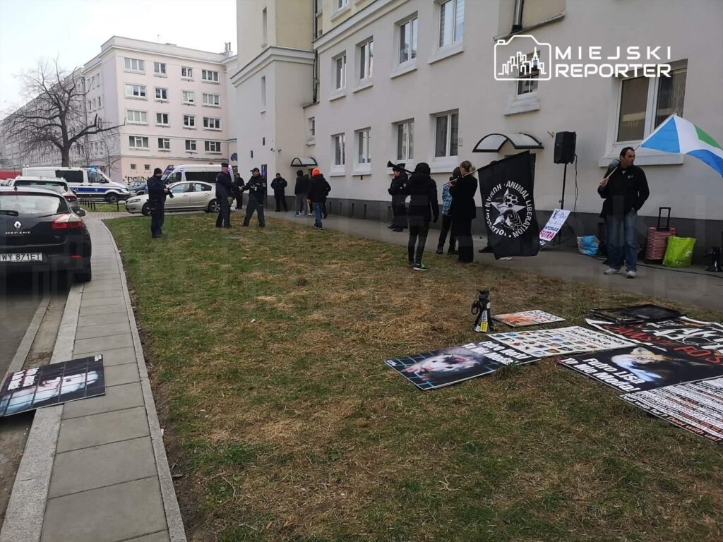 Grupa protestujących z transparentami i flagą wzywającą do ochrony praw zwierząt, stojąca przed budynkiem.