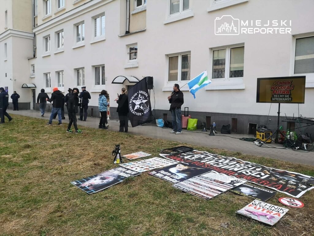 Grupa protestujących z transparentami i flagami stoi przed budynkiem, niektórzy rozmawiają z policją.