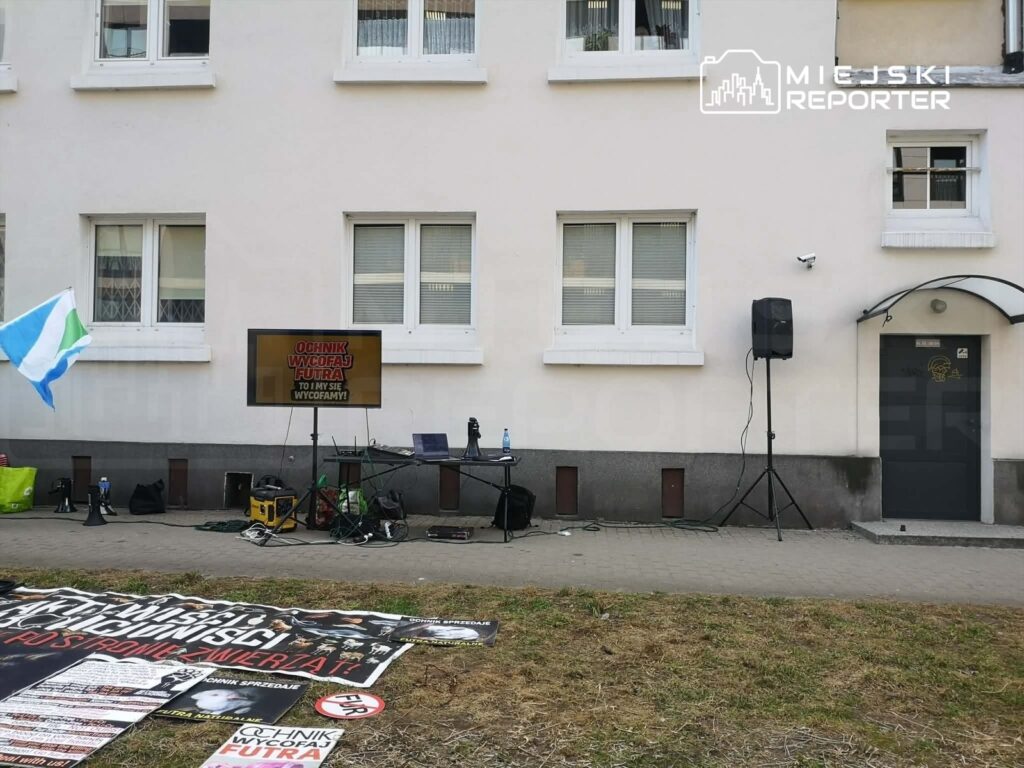 Na chodniku przed budynkiem stoi ekran z napisem "OCHNIK WYCOFAJ FUTURĘ TO INWERSY". Obok znajdują się plakaty oraz sprzęt nagłaśniający. W tle widoczna jest flaga w zielono-niebieskich kolorach.