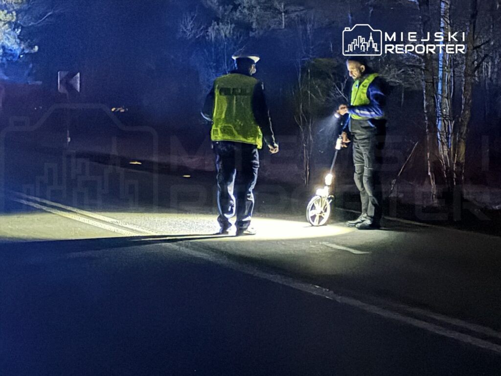 Dwaj policjanci w odblaskowych kamizelkach sprawdzają drogę przy użyciu miernika odległości w ciemnym otoczeniu.