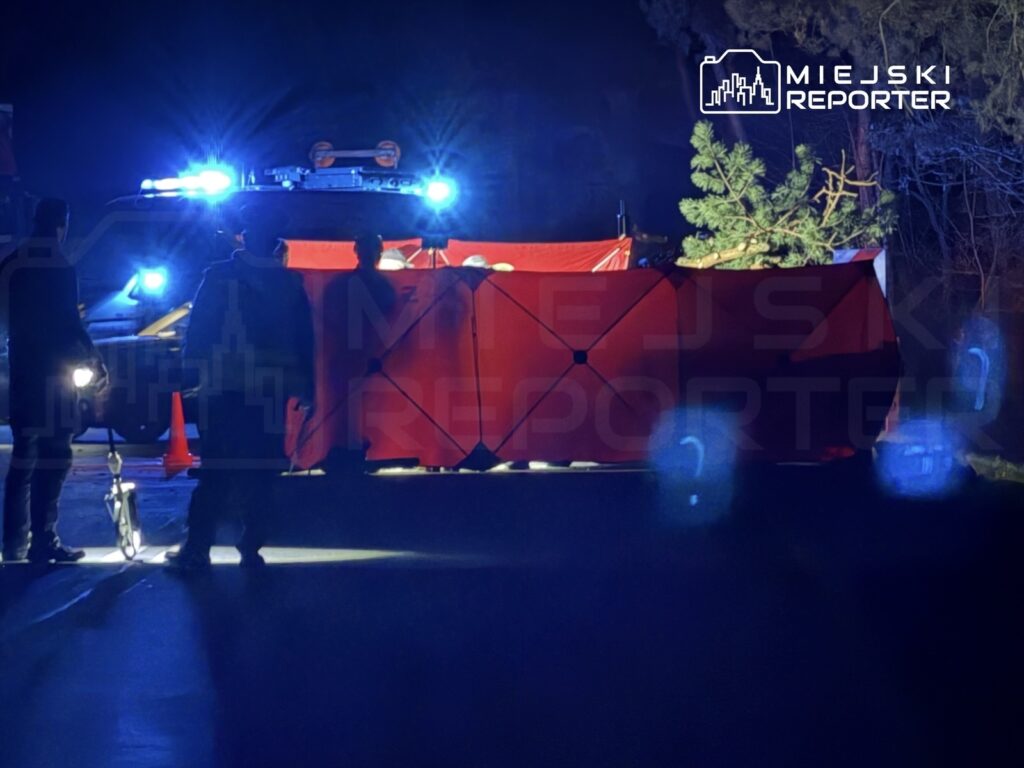 Grupa osób w ciemnym otoczeniu pracuje przy czerwonym namiocie, a w tle świecą niebieskie światła radiowozu.