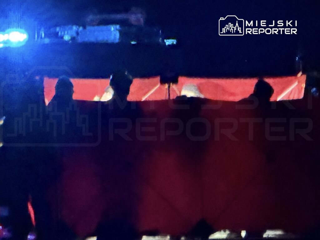 Grupa osób w ciemnych ubraniach stoi za czerwonym materiałem, oświetlona niebieskimi światłami.