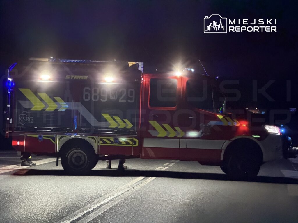 W nocy na drodze stoi wóz strażacki z włączonymi sygnałami świetlnymi, blokując jeden pas ruchu.