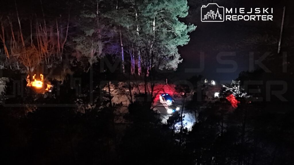 Grupa ratowników z latarkami przeszukuje teren leśny w nocy, obok zaparkowanego pojazdu z włączonymi sygnałami świetlnymi.