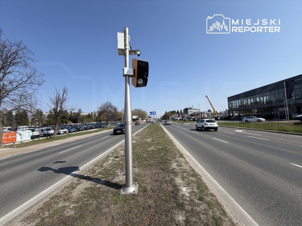 Na drodze znajduje się fotoradar zamontowany na stalowej kolumnie, obok ruchliwej jezdni z samochodami. W tle widoczne są zaparkowane pojazdy oraz nowoczesny budynek.