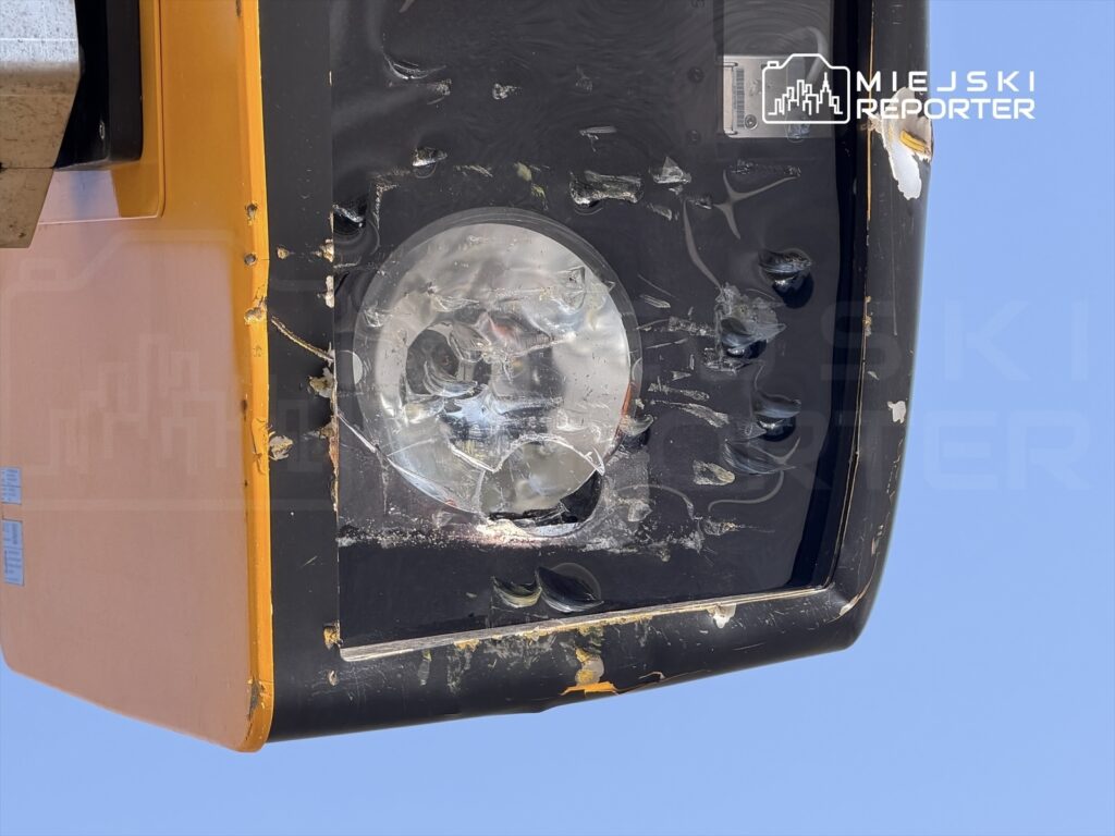 Uszkodzony reflektor w pomarańczowej obudowie zamontowany na urządzeniu budowlanym, w tle błękitne niebo.