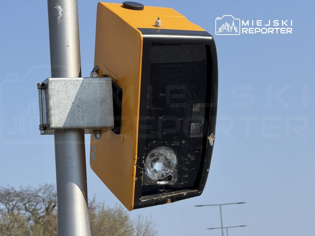 Sygnalizator świetlny w kolorze pomarańczowym zamontowany na stalowym słupie, w tle widoczne niebo i drogi.