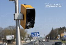 Zniszczony fotoradar zamontowany na słupie przy ruchliwej drodze, w tle widoczne pojazdy i znaki drogowe.