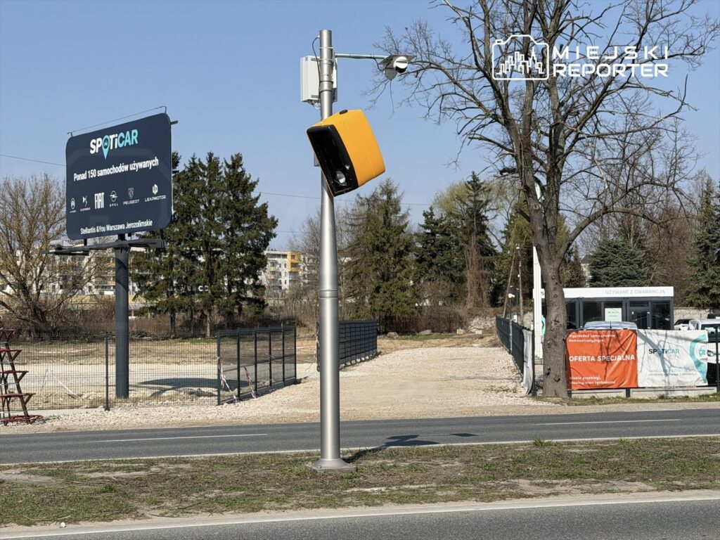 Nowoczesny radar do pomiaru prędkości z żółtą obudową ustawiony przy drodze obok billboardu reklamowego.