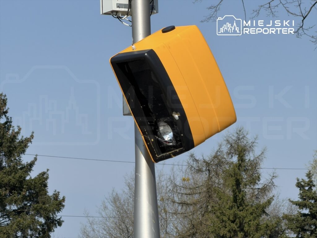Uszkodzony żółty fotoradar zamontowany na słupie w otoczeniu drzew i błękitnego nieba.