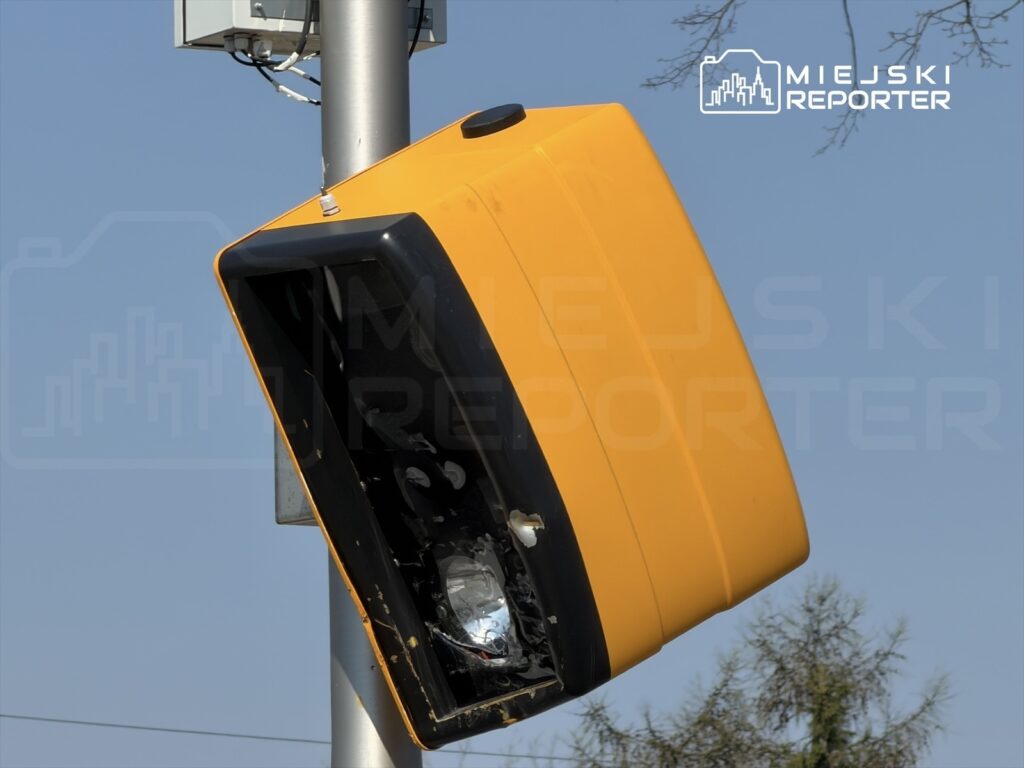 Żółta kamera monitoringu zamontowana na słupie, zwrócona w stronę nieba, w tle jasne niebo i drzewa.