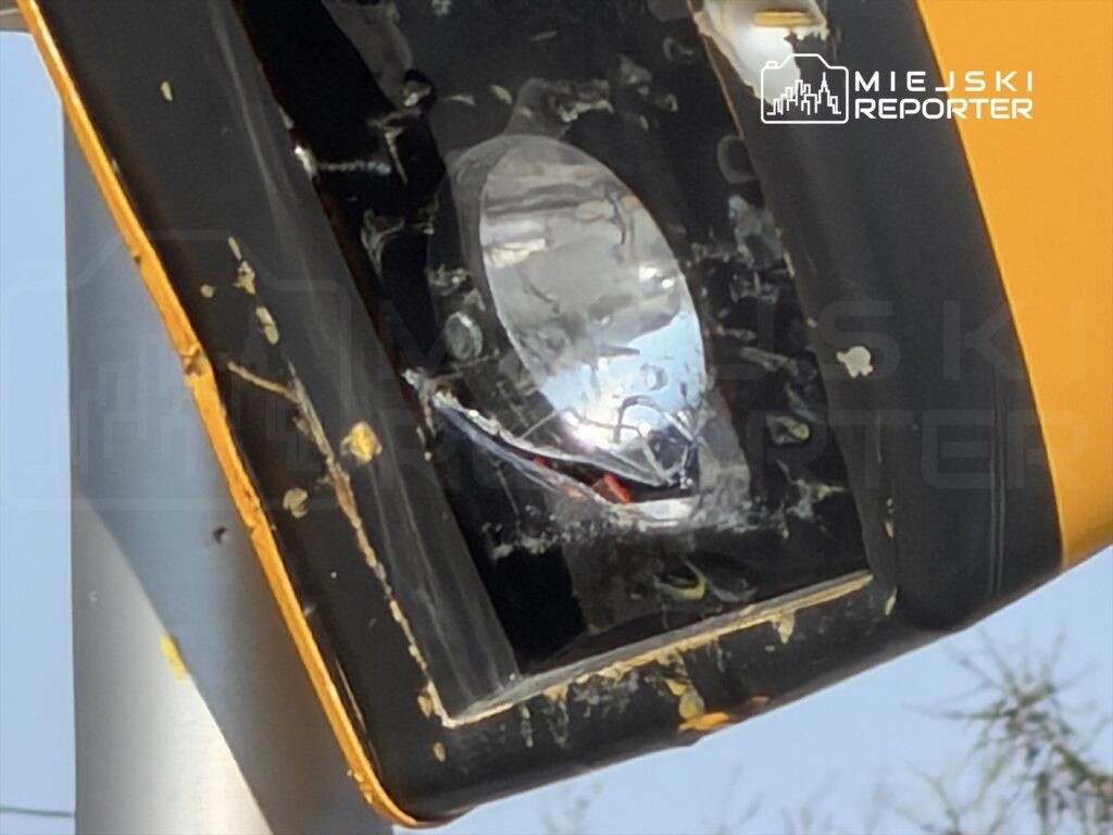 Uszkodzony sygnalizator świetlny z widocznymi rysami i zabrudzeniami, zamontowany na słupie.