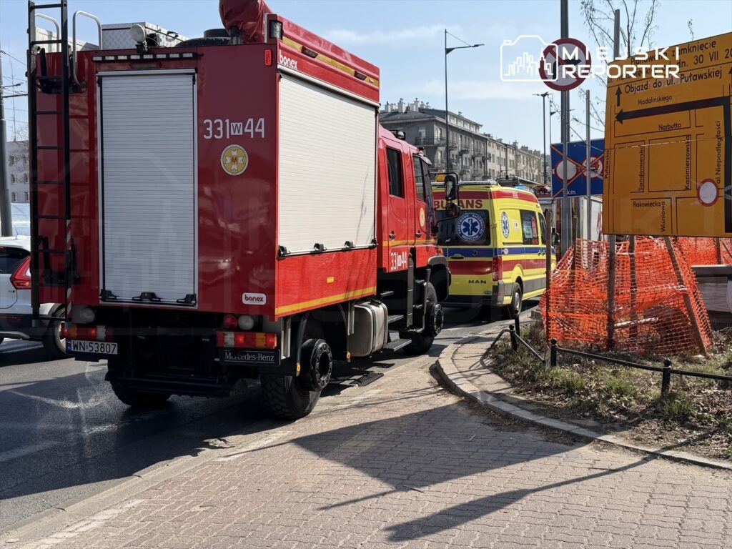 Czerwony wóz strażacki oraz karetka pogotowia zatrzymane na rondzie w miejskim otoczeniu, obok znaków drogowych i ogrodzenia budowy.