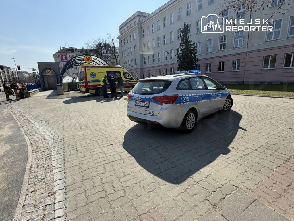 Karetka pogotowia oraz radiowóz policyjny zatrzymane przy wejściu do stacji metra, obok grupy ratowników.