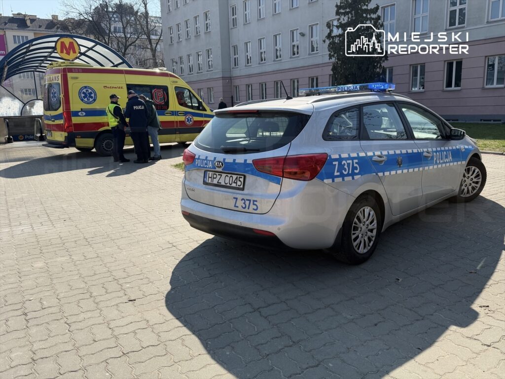 Policjanci rozmawiają z ratownikami medycznymi obok karetki na parkingu przy budynku.